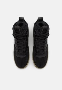 Ett par svarta högskurna sneakers med texturerade tåkuppskärningar, snören och beige räfflade sulor mot en vit bakgrund.