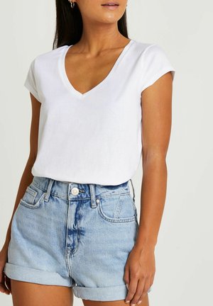 Žena nosí bílý krátký top s výstřihem do V, zastrčený do světle modrých vyhrnutých džínových šortek s vysokým pasem.