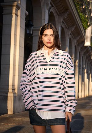 Jeune femme en chemise de rugby rayée avec le texte "Stockholm", chemise blanche à col et jupe en denim, debout dans une arche urbaine baignée de soleil.