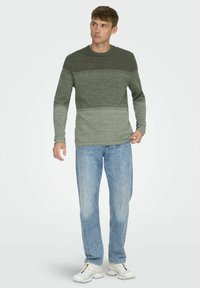Langarmtrui in donkergroene en lichtgroene tinten, gebreide textuur met horizontale strepen, gecombineerd met lichtblauwe jeans en witte sneakers.