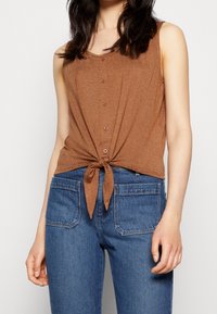 Korte, mouwloze top in bruin met een geknoopt voorpand en knoopdetails; gemaakt van textuurstof met subtiele strepen. Gecombineerd met blauwe denim jeans.