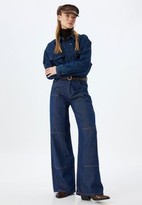 Conjunto de denim que incluye una camisa de botones de manga larga en azul oscuro y pantalones de pierna ancha. Incluye un cinturón y una gorra marrones, con detalles de costura sutiles.