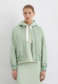 Giacca bomber verde chiaro con finitura lucida, cappuccio, polsini a costine e zip frontale. Indossata sopra un hoodie coordinato.
