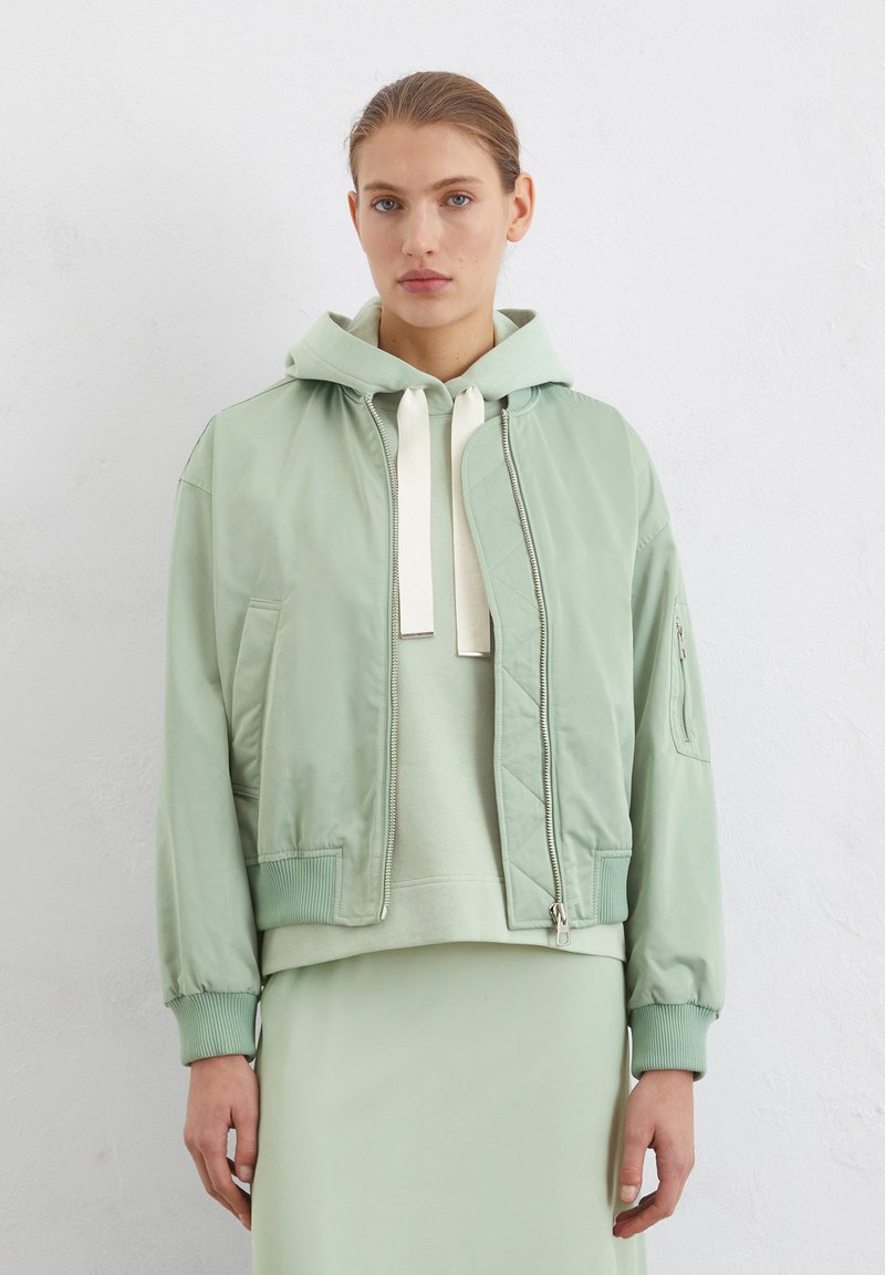 Giacca bomber verde chiaro con finitura lucida, cappuccio, polsini a costine e zip frontale. Indossata sopra un hoodie coordinato.