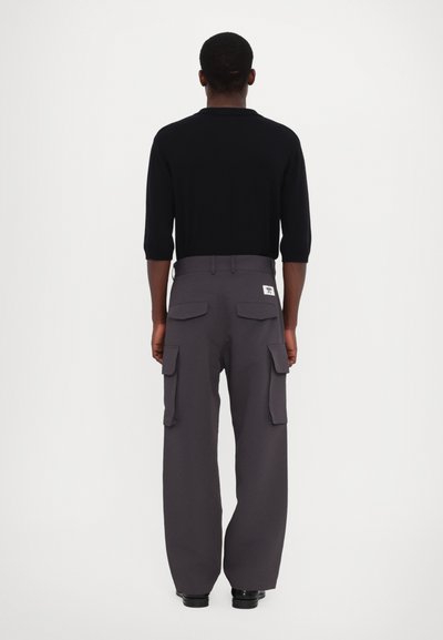 Zwarte polo met korte mouwen, gecombineerd met grijze cargo-broek met wijde pijpen, voorzien van zijzakken en een label op de achterste tailleband.