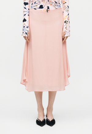 Γυναίκα που φοράει μια ροζ ανοιχτή midi φούστα με ασύμμετρα πλαϊνά, συνδυασμένη με μπλούζα με μακριά μανίκια και λουλούδια, και μαύρα παπούτσια με μυτερή μύτη.