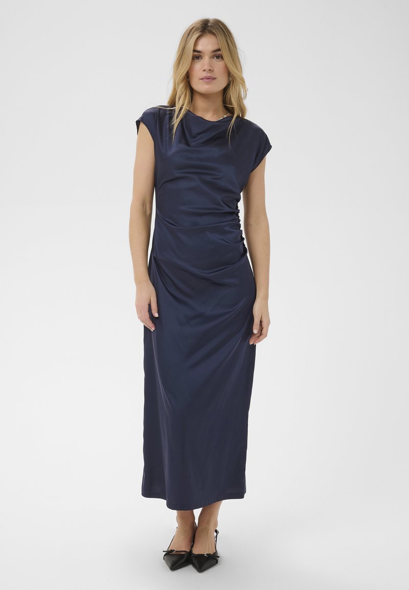 Femme portant une robe longue sans manches en satin bleu marine et des escarpins noirs à bout pointu, debout devant un fond blanc uni.