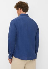 Blue linen long-sleeve shirt with button cuffs and a back yoke. The fabric has a textured appearance, with a relaxed fit and curved hemline.