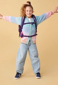 Cardigan multicolore con strisce rosa e blu, indossato sopra una camicia bianca. Jeans leggeri azzurri e sneakers navy, in spalla uno zaino viola.