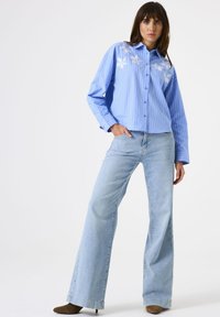 Woman wearing a blue striped button-up shirt with white floral embroidery and light blue wide-leg jeans, posing with one hand in pocket.