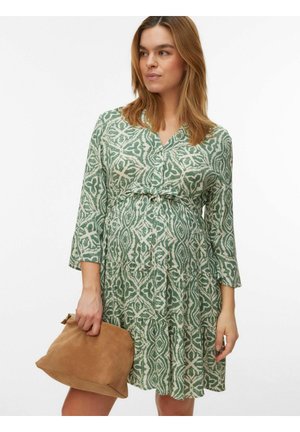 Rase naine, kes kannab rohevalget mustrilist kleiti kolmeveerandvarrukatega, hoides pruuni seemisnahast käekotti.