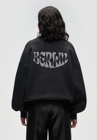 Black oversized sweatshirt with a matte finish, featuring the word "BERLIN" in bold, light gray lettering on the back. Smooth texture.