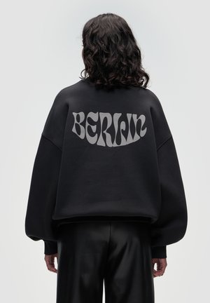 Czarna, oversize bluza z matowym wykończeniem, z napisem "BERLIN" w odważnym, jasnoszarym kolorze na plecach. Gładka struktura.