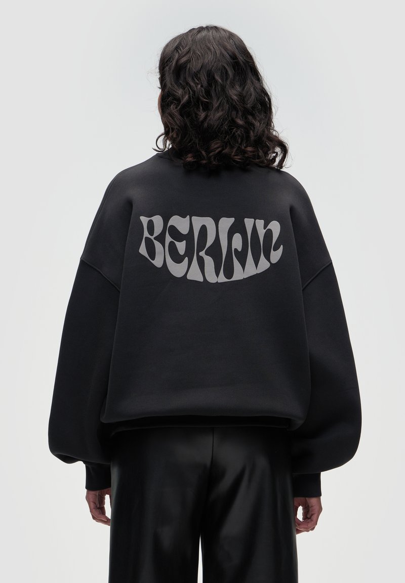 Black oversized sweatshirt with a matte finish, featuring the word "BERLIN" in bold, light gray lettering on the back. Smooth texture.