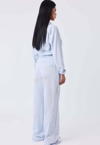Vrouw met lang zwart haar, gekleed in een lichtblauwe hoodie en losse bijpassende broek, staande zijwaarts tegen een effen witte achtergrond.
