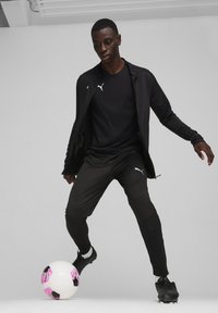 Veste et pantalon de sport noirs avec des accents réfléchissants, associés à des chaussures de football noires. Joueur positionné pour frapper un ballon de football rose et noir.