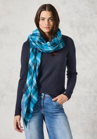 Écharpe à carreaux bleue et turquoise en tissu doux, drapée autour du cou. Portée avec un pull sombre et un jean bleu clair.