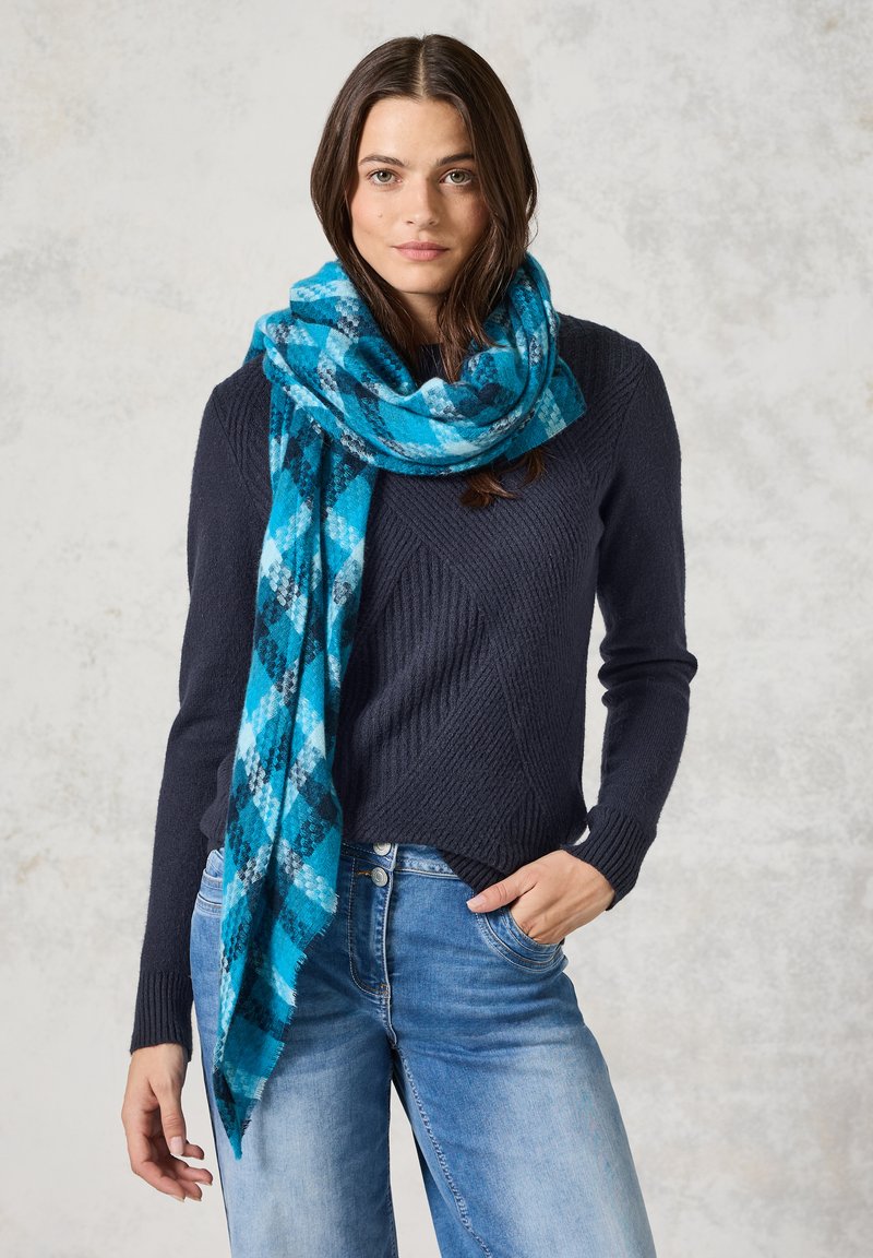 Écharpe à carreaux bleue et turquoise en tissu doux, drapée autour du cou. Portée avec un pull sombre et un jean bleu clair.