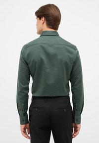 Uomo che indossa una camicia a maniche lunghe verde scuro infilata nei pantaloni eleganti neri, visto di spalle su uno sfondo bianco semplice.