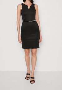 Robe noire sans manches avec un décolleté en V et une silhouette cintrée, ornée d'une fine ceinture blanche. Tissu lisse sans motifs visibles.