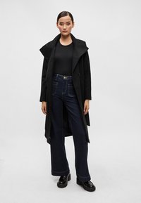 Hosszú fekete kabát széles hajtókákkal, fekete, testhezálló felsővel és magas derekú, sötét farmer bőrnadrággal párosítva. A platform cipők egészítik ki a megjelenést.