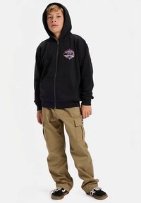 Camisola com capuz preta com fecho de correr frontal, bolsos à frente e logótipo multicolorido, combinada com calças cargo bege com bolsos laterais e ténis pretos.