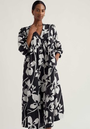 Kobieta w długiej czarnej sukni z dużym białym kwiatowym wzorem, z dekoltem w kształcie litery V i bufkami na długich rękawach, pozująca z jedną ręką przy szyi.