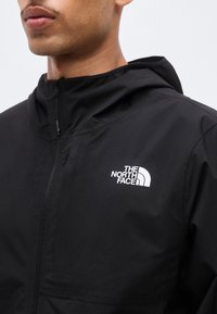 Zbliżenie na mężczyznę noszącego czarną kurtkę zapinaną na zamek z kapturem i białym logo The North Face na piersi.
