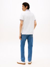 Weißes Polohemd mit gestreiftem Kragen, kombiniert mit blauen Jeans. Modell blickt rückwärts und trägt weiße Sneakers mit Gummisohle.