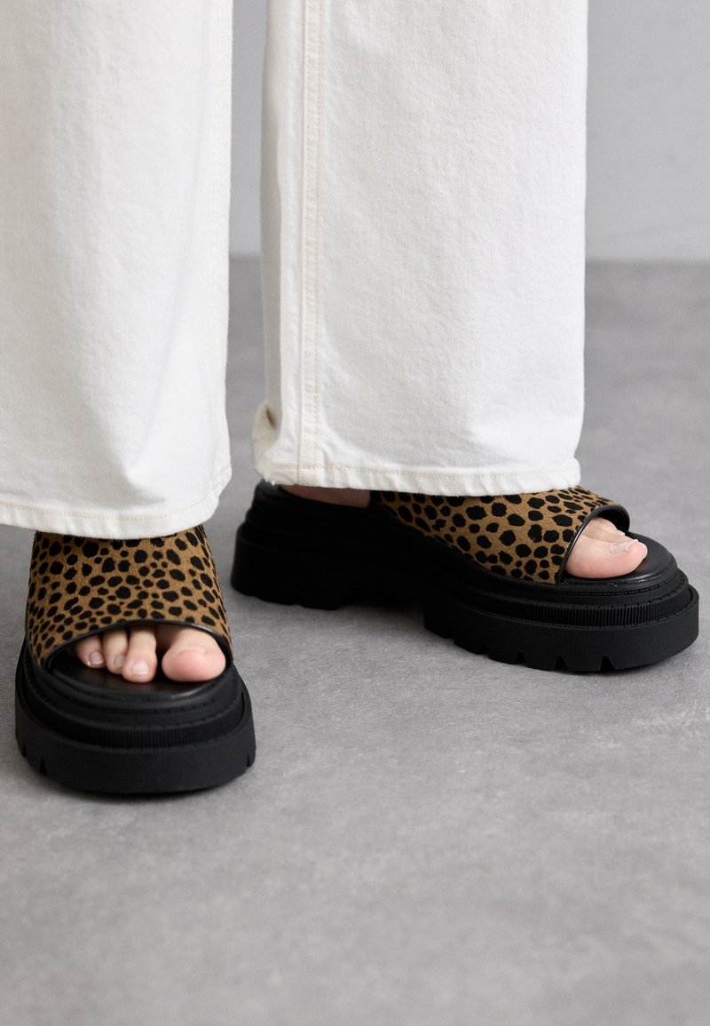 Pieds portant un pantalon blanc à jambes larges et des sandales à plateforme à orteils ouverts avec des semelles noires et des lanières imprimées léopard marron sur un sol gris.