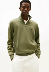 Modèle masculin portant un sweat à col zippé olive sur une chemise blanche, les mains dans les poches, se tenant contre un fond blanc uni.