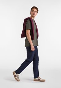 Camiseta verde oliva, sudadera marrón drapeada sobre los hombros, pantalones azul marino y zapatillas beige con acentos marrones.