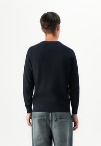 Bărbat întors cu spatele, îmbrăcat cu un pulover lung cu mâneci lungi, texturat, de culoare bleumarin închis și blugi din denim albastru, largi, cu buzunare mari pe spate.