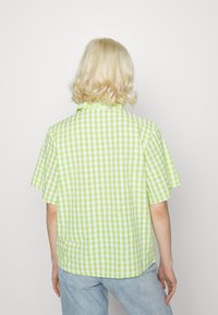 Camisa de manga curta, em padrão xadrez verde e branco, com um corte descontraído e colarinho, apresentando uma bainha reta e sem fechos visíveis.