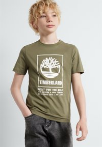 Olijfgroene katoenen T-shirt met een witte afbeelding van een boom en tekst. Ronde hals, korte mouwen, relaxte pasvorm, gecombineerd met grijze denim broek.