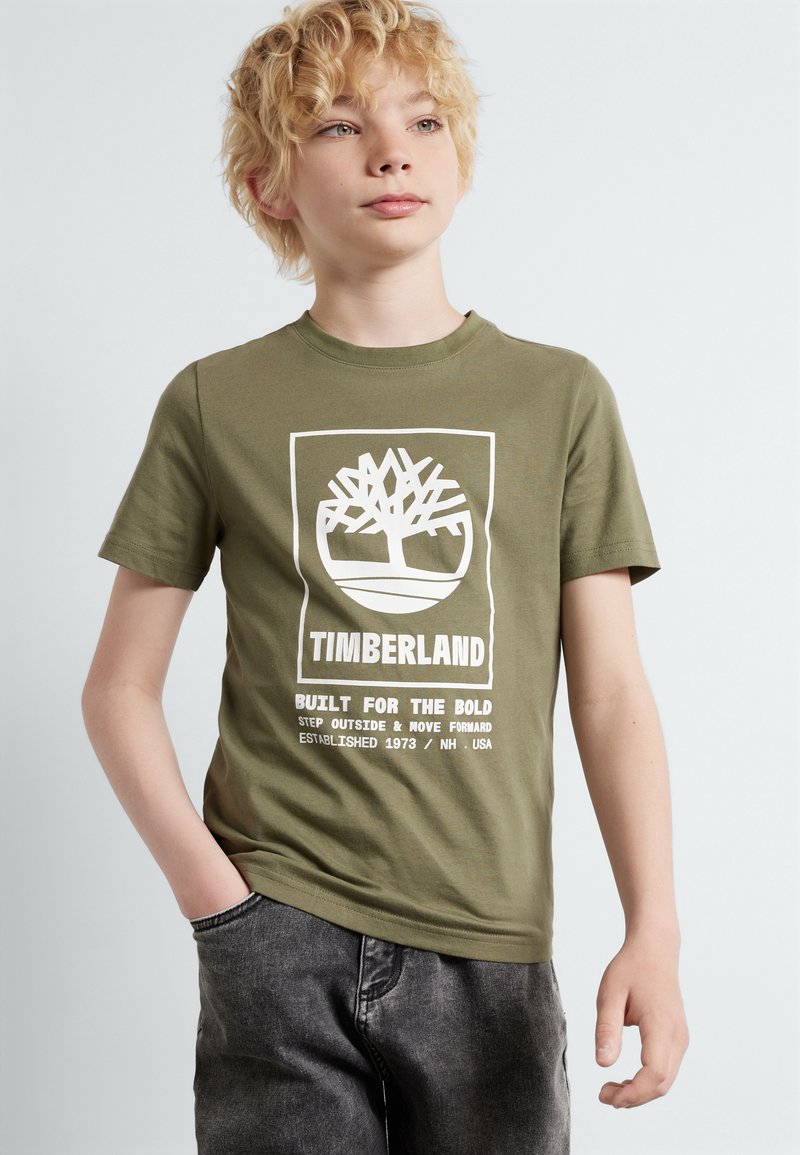 Olijfgroene katoenen T-shirt met een witte afbeelding van een boom en tekst. Ronde hals, korte mouwen, relaxte pasvorm, gecombineerd met grijze denim broek.