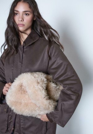 Young woman with long dark hair wearing a brown coat holds a large beige fluffy bag against a plain light background.