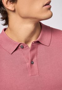 Polo shirt in morbido tessuto rosa con una finitura testurizzata, con colletto e due bottoni neri al collo. Vista ravvicinata del colletto.