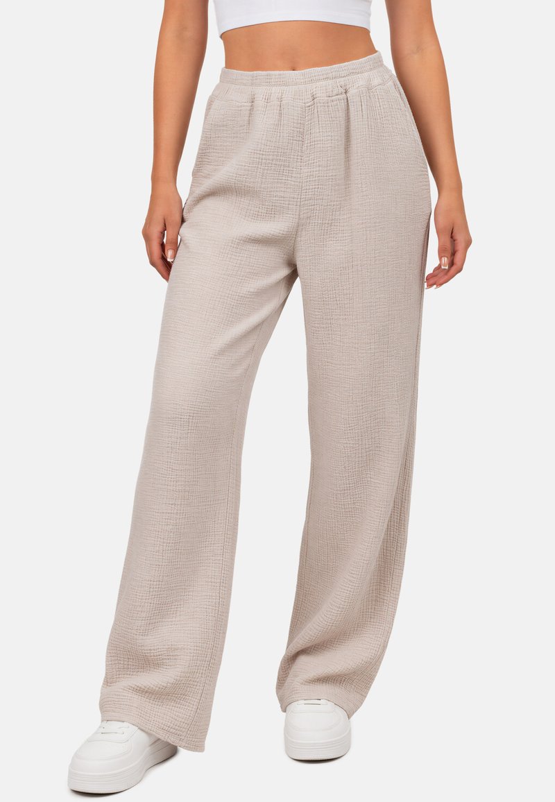 Femme portant un pantalon beige ample texturé et des baskets blanches avec un top blanc court sur un fond uni.
