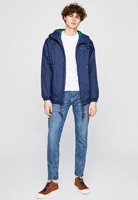 Giacca leggera blu navy con dettagli verdi, cappuccio con cordino, polsini elasticizzati e due tasche laterali, abbinata a jeans blu e scarpe da ginnastica marroni.