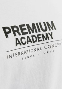 Balta medvilninė marškinėlių su drąsiai juodu tekstu: "PREMIUM ACADEMY" ir "INTERNATIONAL CONCEPT SINCE - 1990" paprasto dizaino.