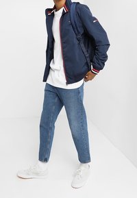 Osoba oblečená v námořnickém saku s bílým a červeným lemem, bílém tričku, modrých džínách, bílých ponožkách, bílých teniskách a nese námořnický batoh.