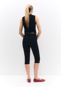 Černý top bez rukávů s detailním vázáním v pase a černými legínami, doplněný červenými balerínami, z pohledu zezadu na bílém pozadí.