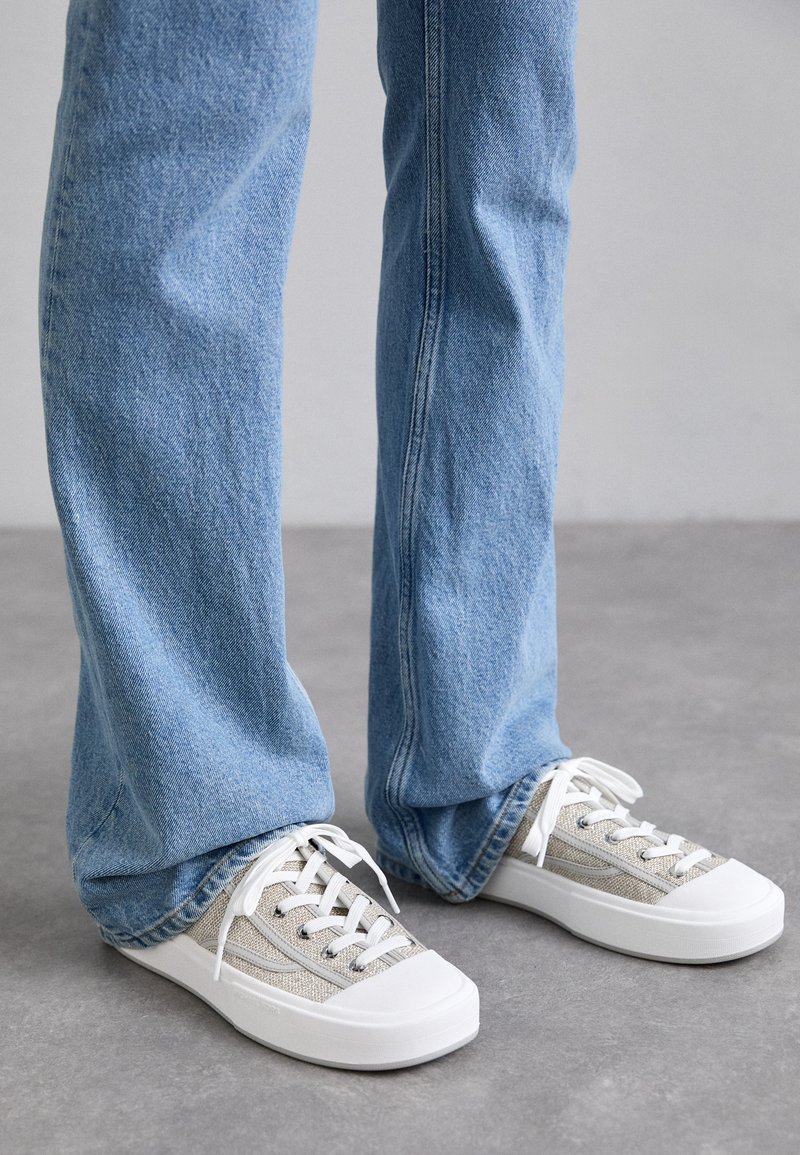 Beige lerretsko med hvite gummisåler og snøring, kombinert med lyseblå jeans. Synlig søm og en avslappet passform.