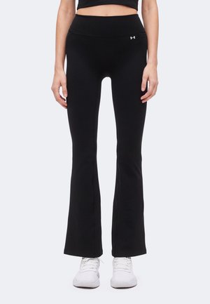 Kvinde iført sorte tætsiddende flare leggings med Under Armour-logo og hvide sportssko står mod en ensfarvet baggrund.