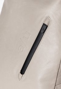 Beige leren rugzak met een gladde textuur, een geperst logo en een zwarte ritszak aan de voorkant voor veilige opslag.
