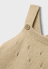 Beige gestricktes Kleidungsstück mit einem strukturierten Muster und einem einzelnen Holzknopf am Schulterriemen. Weicher, leichter Stoff.