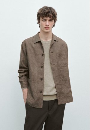 Jeune homme aux cheveux bouclés portant une veste marron à boutons sur un pull beige et un pantalon foncé, debout devant un fond clair uni.