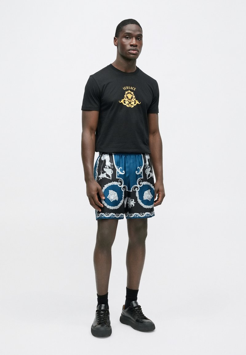 Sort bomulds-T-shirt med guld "Versace"-logo. Blå shorts med detaljerede barokmønstre i sort og sølv. Sorte sneakers fuldender outfittet.