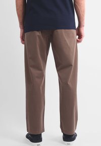 Pantalon en coton marron à coupe droite, avec une texture lisse et des plis subtils. Porté avec des chaussures foncées ; étiquette d'entretien visible.